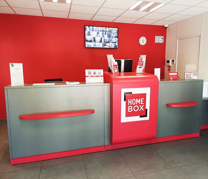 Comptoir d'accueil rouge et gris avec logo HOMEBOX et écran de surveillance du centre HOMEBOX Les Herbiers