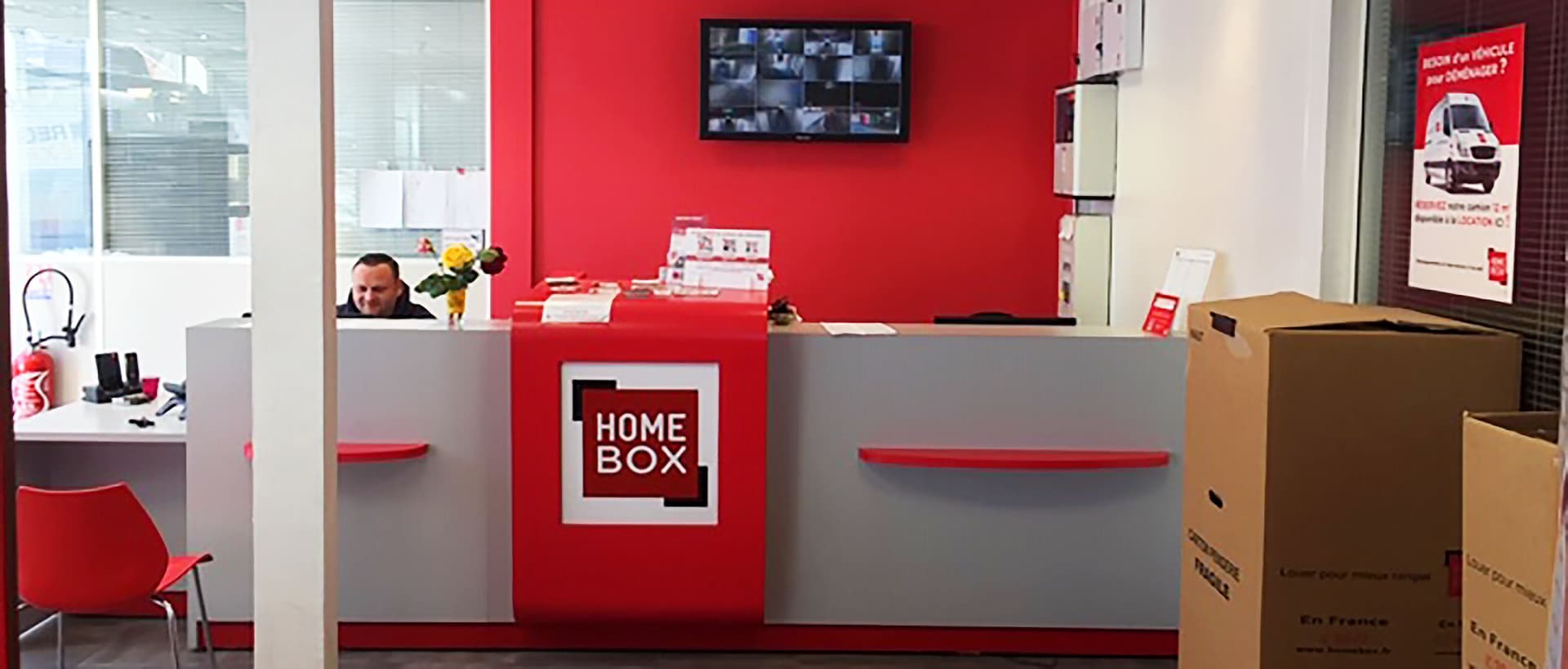 Conseiller au comptoir d'accueil rouge avec écran de vidéosurveillance et cartons, HOMEBOX Rouen