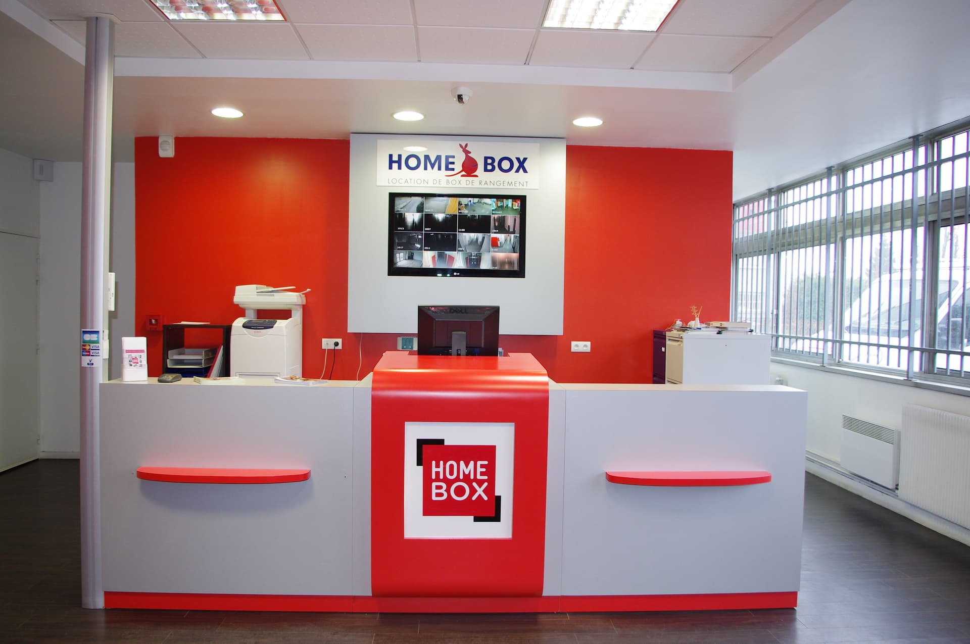 Comptoir d'accueil rouge et blanc avec écran de vidéosurveillance du centre HOMEBOX Maurepas