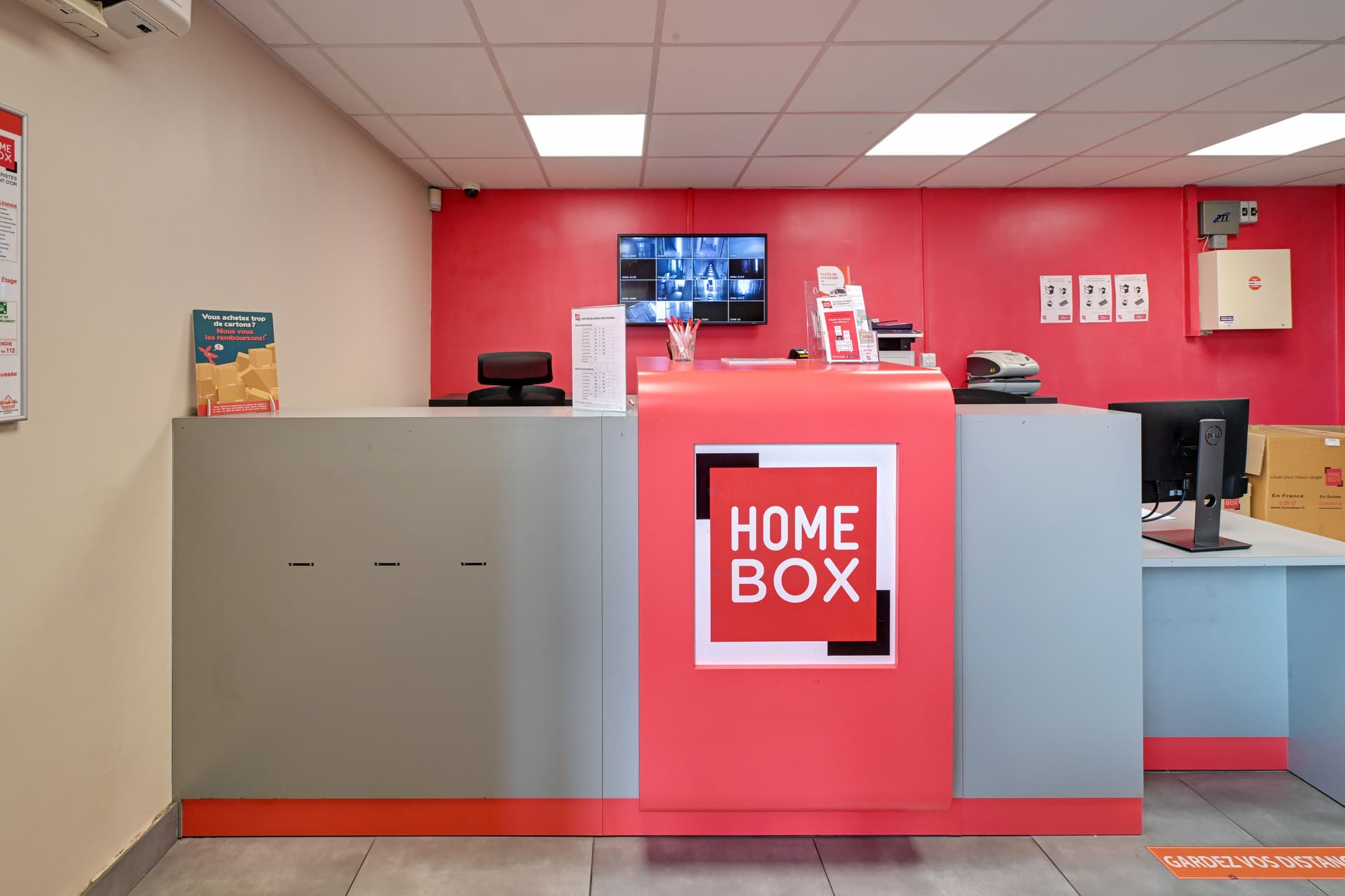 Comptoir d'accueil rouge avec logo HOMEBOX et écrans de vidéosurveillance du centre HOMEBOX Lyon Ouest