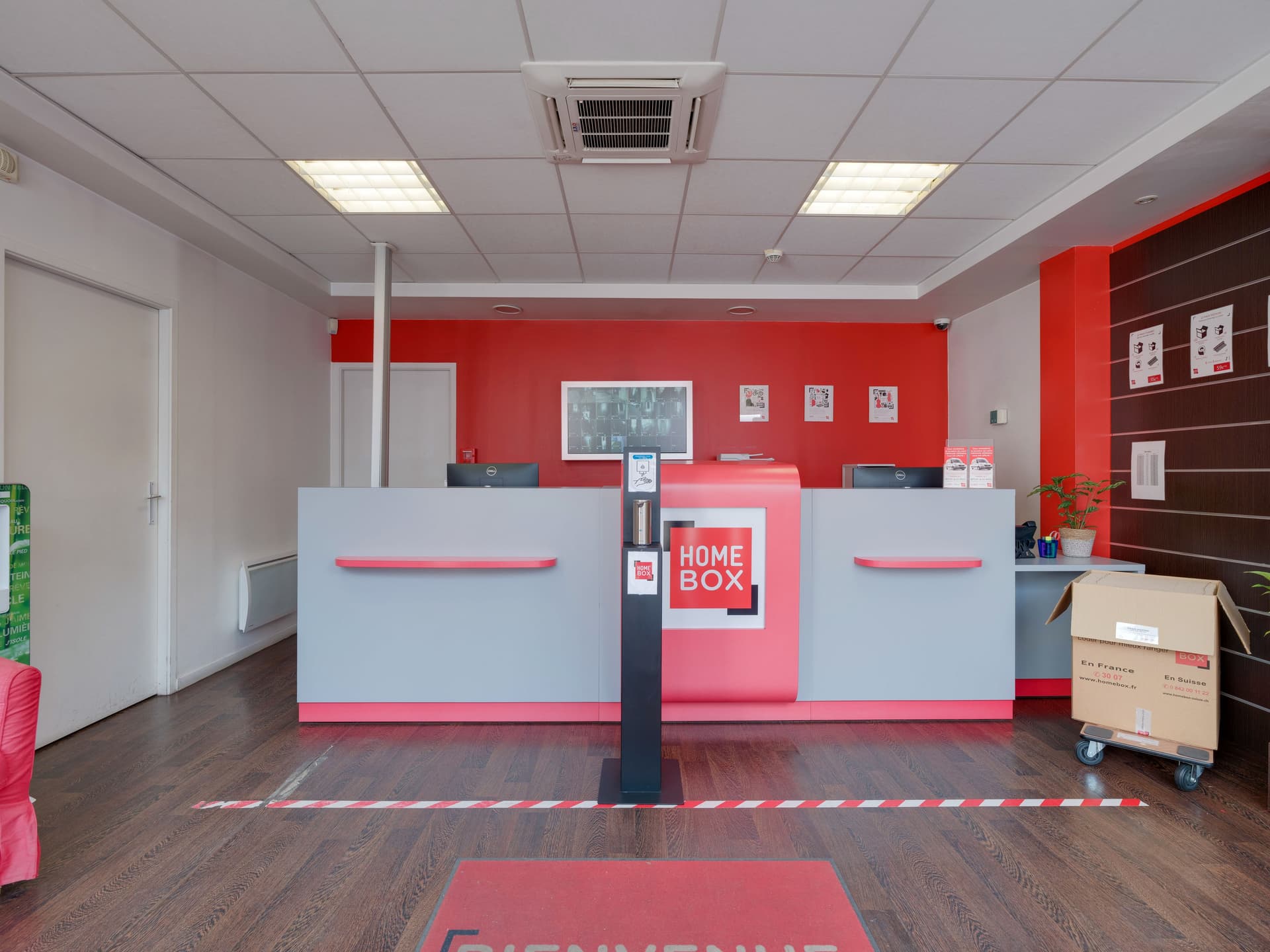 Comptoir d’accueil du centre HOMEBOX Annecy avec mur rouge et écran mural.
