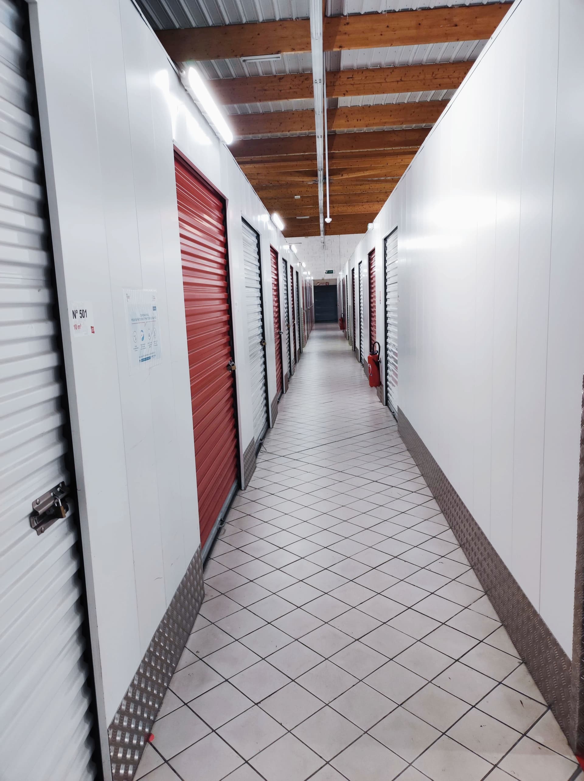 Couloir intérieur carrelé bordé de box de stockage à portes rouges sous charpente bois du centre HOMEBOX Périgueux