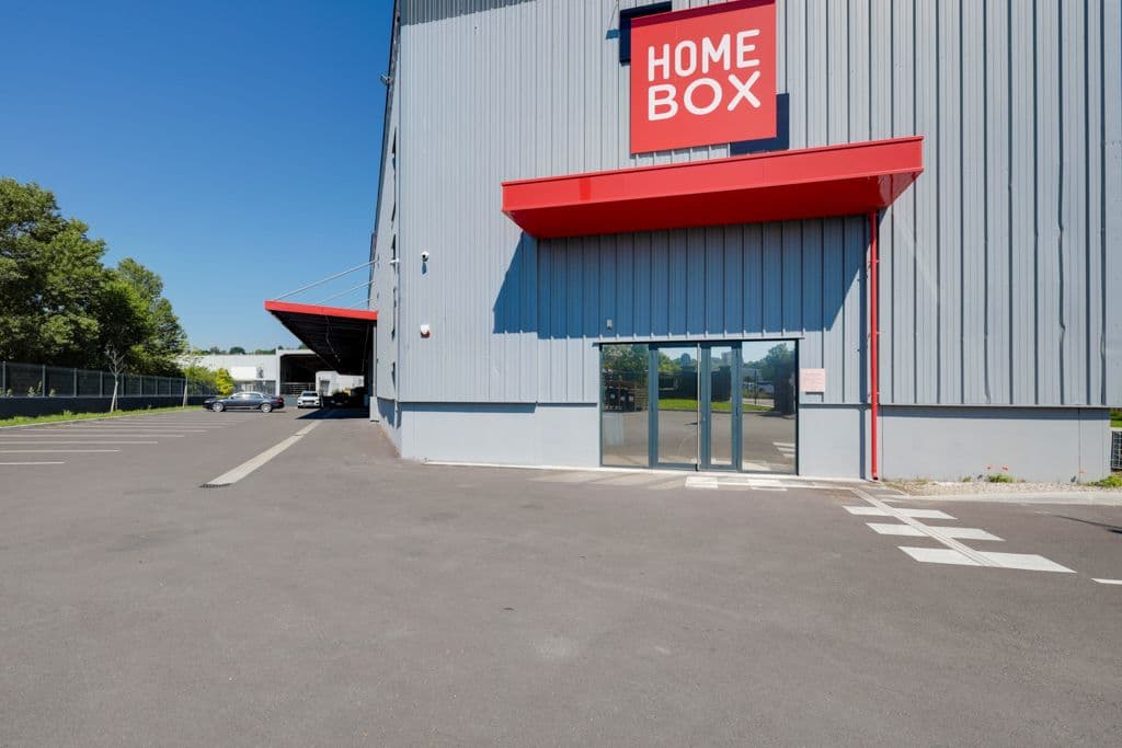 Façade grise avec auvent rouge et entrée vitrée du centre vue depuis le parking, HOMEBOX Saint-Ouen-l'Aumône, 1 Rue des Préaux à Saint-Ouen-l'Aumône