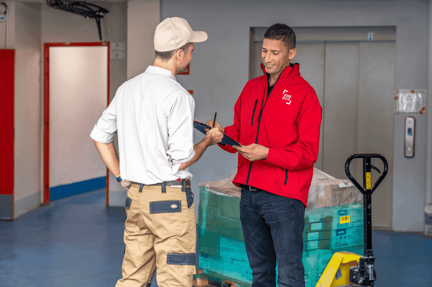 Échange entre un conseiller HOMEBOX et un client professionnel devant un box de stockage, avec palettes de marchandises et transpalette.